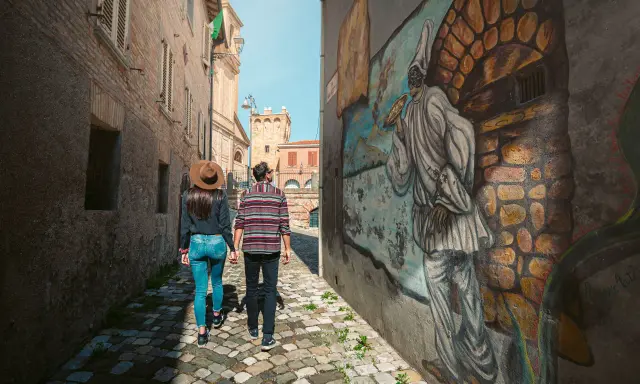 Coppia passeggia in un vicolo di Saludecio, ammirando un murale. Scopri cosa vedere in questo borgo incantevole.