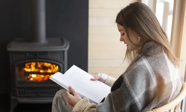 Une femme lit un livre près d'un poêle à granulés sans électricité. Avis : chaleur réconfortante et ambiance cosy.