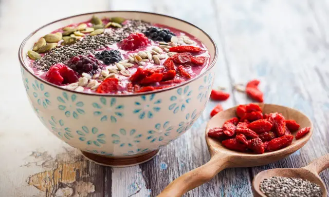 Miseczka smoothie z owocami, nasionami chia, pestkami dyni i słonecznika. Obok łyżka z jagodami goji. Nasiona chia jak jeść? Dodaj do smoothie!