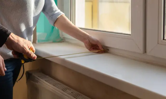 Dwie osoby mierzą taśmą zwymiarową parapet wewnętrzny przy oknie.