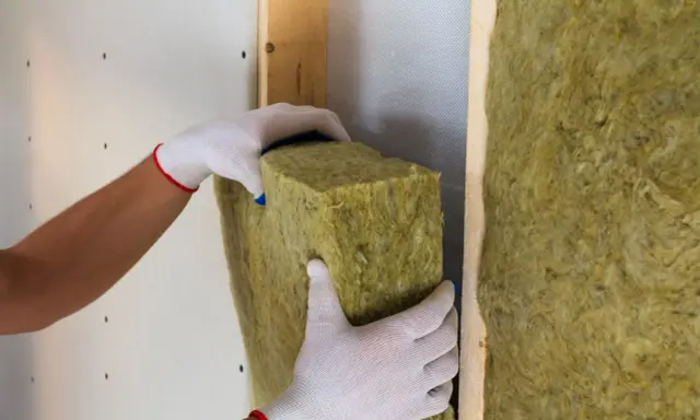 Manos enguantadas colocan aislamiento de lana de roca para como aislar una pared, creando un hogar más cálido y eficiente.