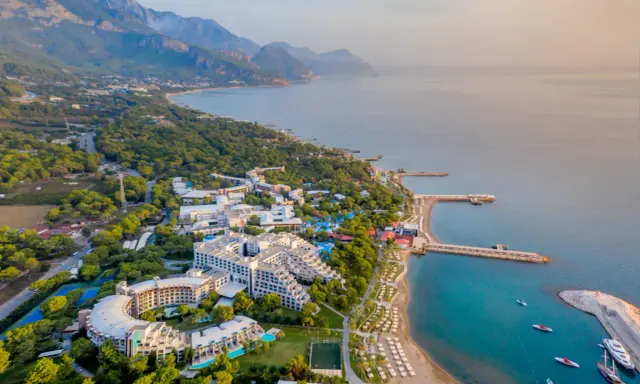 Widok z lotu ptaka na Rixos Sungate, luksusowy kurort nad turkusowym morzem, otoczony zielenią i górami.