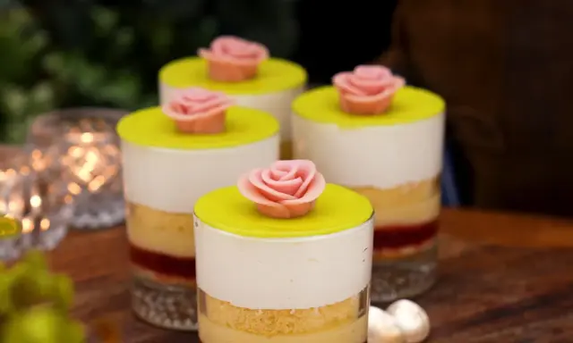 Fyra små prinsesstårta i glas, dekorerade med rosa marsipanrosor och gul marsipanlock. Vackert uppdukat med ljus i bakgrunden.