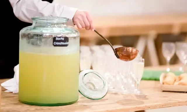 Une louche sert une soupe champenoise sans alcool dans un grand pichet en verre. Des verres à pied et des amuse-bouches sont disposés sur la table.