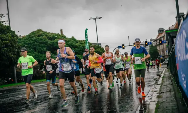 Uczestnicy maratonu biegną w deszczu po mokrej nawierzchni. Widać ich skupienie i determinację mimo niesprzyjającej pogody.