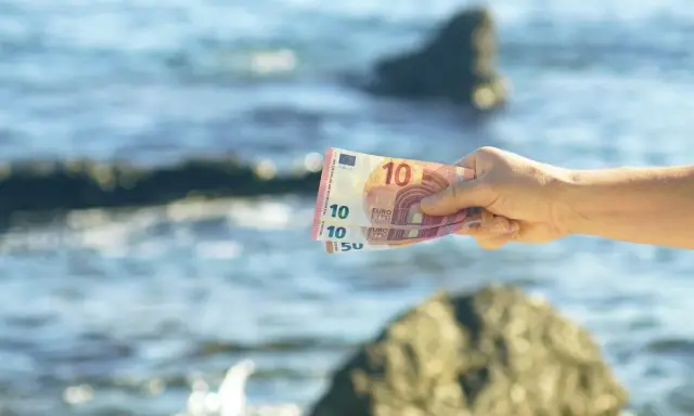 Dłoń trzyma banknoty euro na tle morza. W Czarnogórze obowiązuje euro, więc te pieniądze przydadzą się na wakacjach.