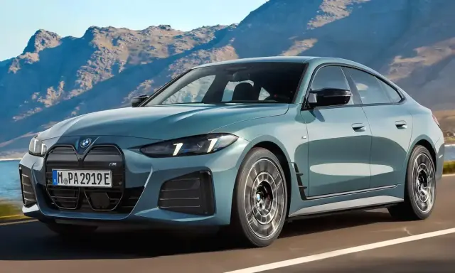 Der neue BMW i4 Facelift in einem eleganten Blau fährt auf einer Küstenstraße vor einer Bergkulisse.