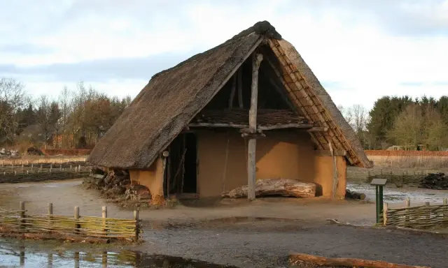 Ein rustikales Bauernhaus aus dem Mittelalter mit Strohdach und Lehmwänden, umgeben von Holzzäunen und Bäumen.