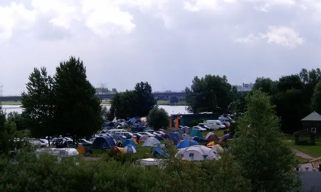 Rozległy kamping z mnóstwem namiotów i samochodów, nad wodą, pod pochmurnym niebem.