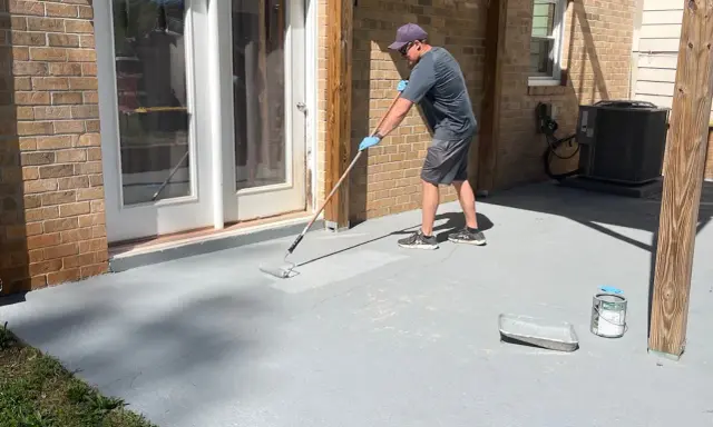 Un homme applique une peinture sur mur béton extérieur avec un rouleau. Le sol gris est en cours de rénovation.