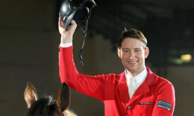 Reiter Franke Sloothaak hält stolz seinen Helm hoch, trägt eine rote Reitjacke und sitzt auf einem braunen Pferd.