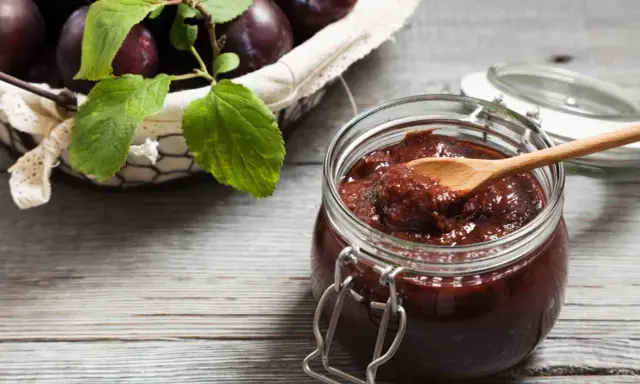 Słoik pełen domowych powideł śliwkowych z czekoladą, obok koszyk z dojrzałymi śliwkami.