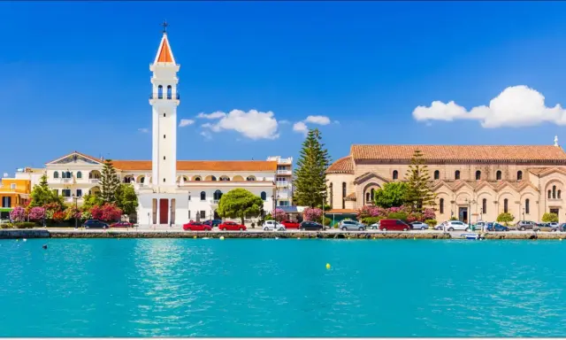 Widok na miasto Zakynthos z turkusową wodą i budynkami, w tym kościołem z charakterystyczną wieżą.