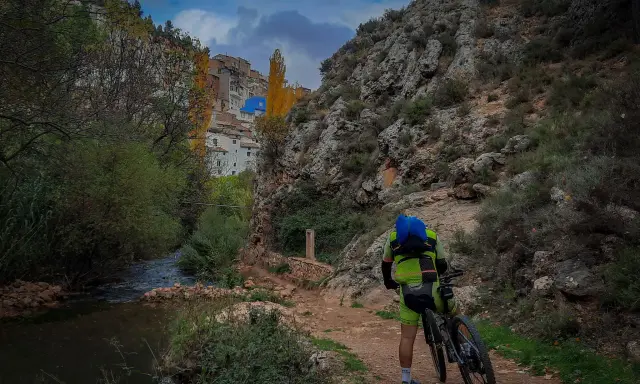 Ciclista en un sendero junto a un río, con un pueblo en la ladera. Perfectas rutas senderismo Albacete.