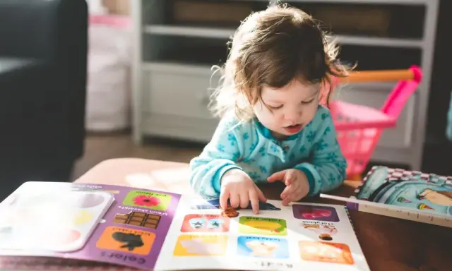 Mała dziewczynka z niepełnosprawnością intelektualną z zaangażowaniem przegląda książeczkę z obrazkami, wskazując paluszkiem na kolorowe ilustracje.