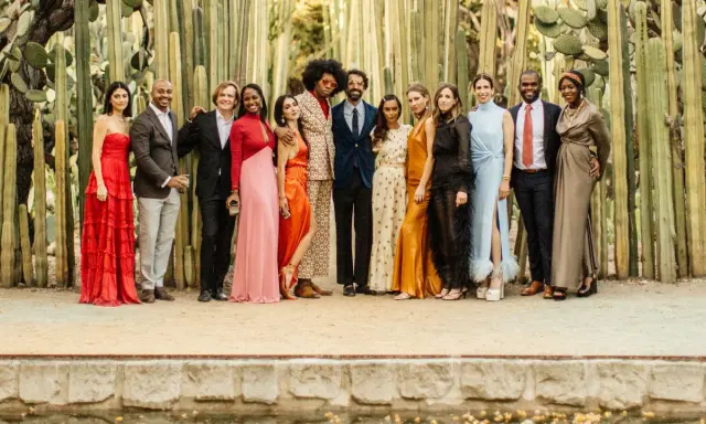 A diverse group of people pose in front of tall cacti, showcasing a vibrant summer color palette with dresses in red, pink, orange, and gold, alongside suits in blues and browns.