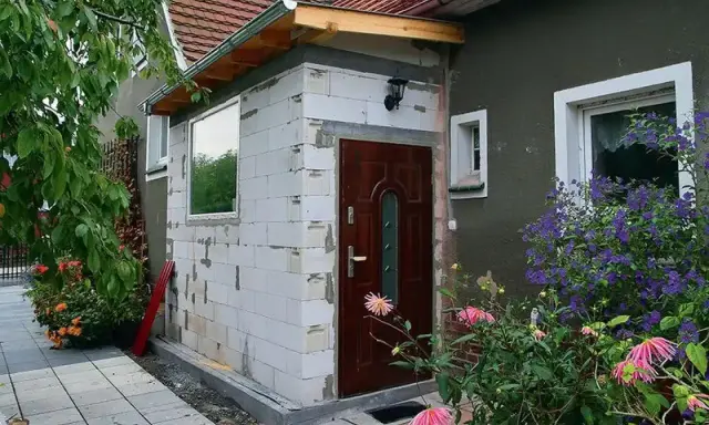 Budowa nowego przedsionka do domu bez pozwolenia. Białe pustaki, brązowe drzwi i okno czekają na wykończenie.