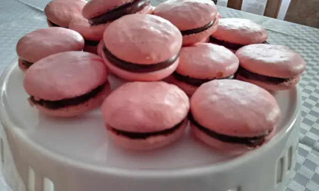Różowe makaroniki na bezie włoskiej z czekoladowym nadzieniem, ułożone na białej paterze.