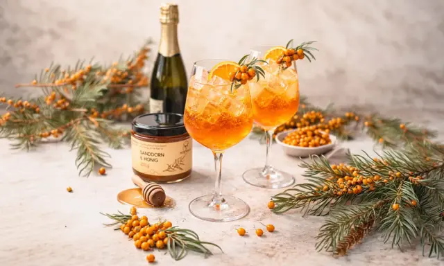 Zwei erfrischende Sanddorn Spritz Cocktails mit Orangenscheiben und Sanddornzweigen, daneben eine Flasche Sekt und ein Glas Sanddorn & Honig.