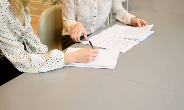 Dwie kobiety omawiają umowę o świadczenie usług. Jedna pisze notatki, druga przegląda dokumenty.