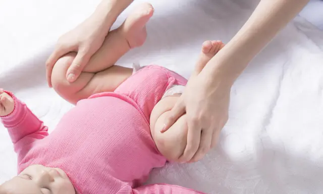 Delikatne ruchy dłoni pomagają w diagnostyce dysplazji stawu biodrowego u niemowlaka w różowym body.
