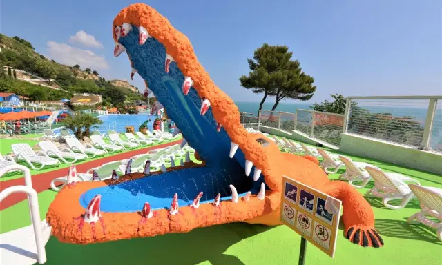 Un gigantesco coccodrillo arancione con la bocca spalancata, un divertente scivolo acquatico, si affaccia su una piscina e lettini bianchi, con una rigogliosa riva verde fermo sullo sfondo.