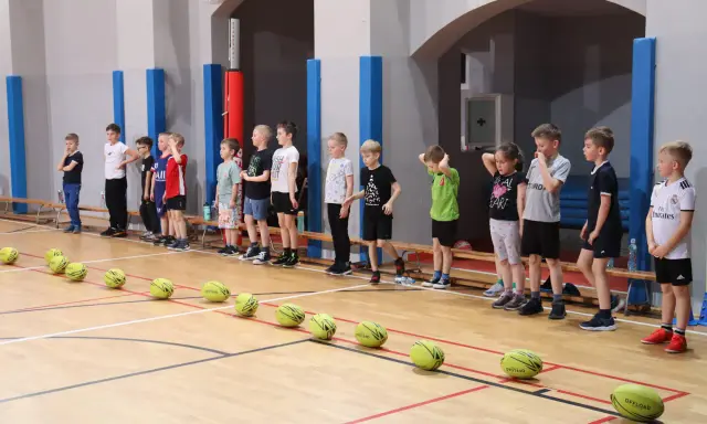 Dzieci w sali gimnastycznej czekają na rozpoczęcie zabaw ruchowych. Przed nimi leżą żółte piłki.