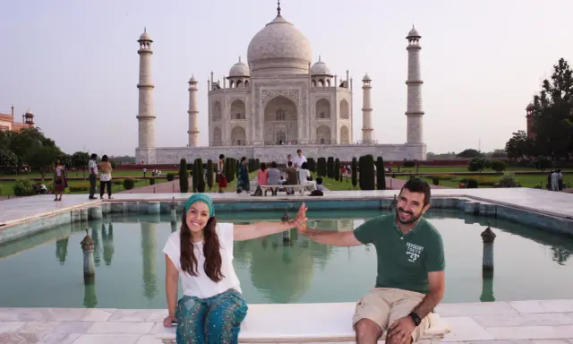 Pareja sonriente en la India, frente al Taj Mahal, con niños y visitantes al fondo.