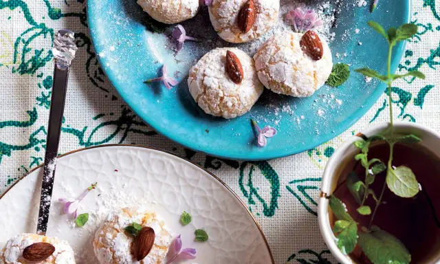 Biscotti al cocco spolverati di zucchero a velo, guarniti con mandorle e fiori viola, accanto a una tazza di tè alla menta.