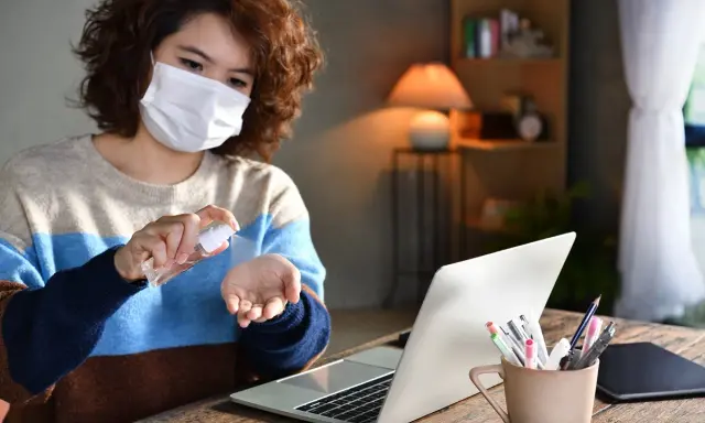 Kobieta w masce dezynfekuje dłonie płynem ze sprayu przy laptopie.