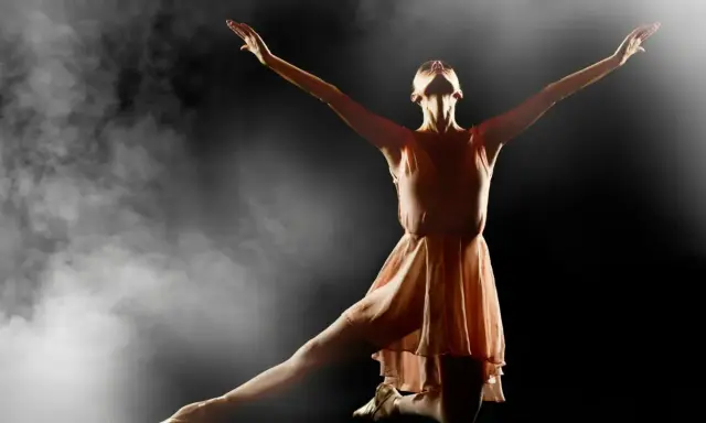 En ballerina i en ljusrosa klänning utför en graciös rörelse i klassisk balett, omgiven av dramatisk rök och ljus.