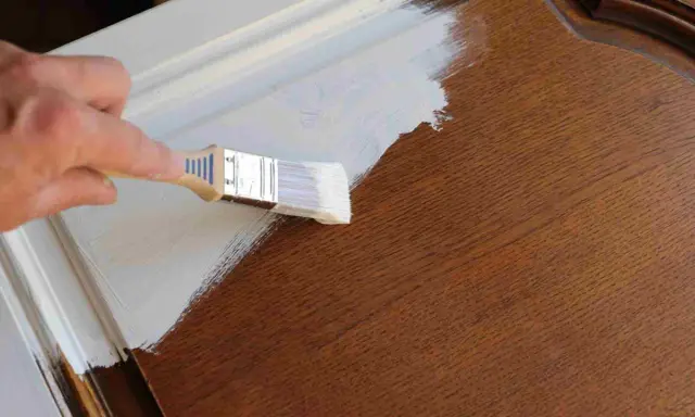 Mano pintando una puerta de madera con brocha blanca. Guía para como pintar puertas de madera.