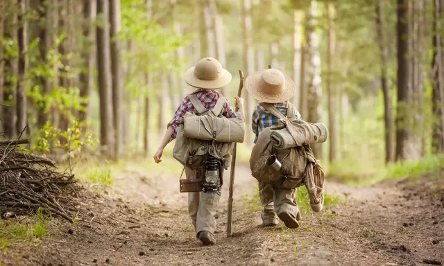 Dwoje dzieci w kapeluszach i z plecakami wyrusza na leśną ścieżkę. To przygoda i nauka survivalu dla dzieci.