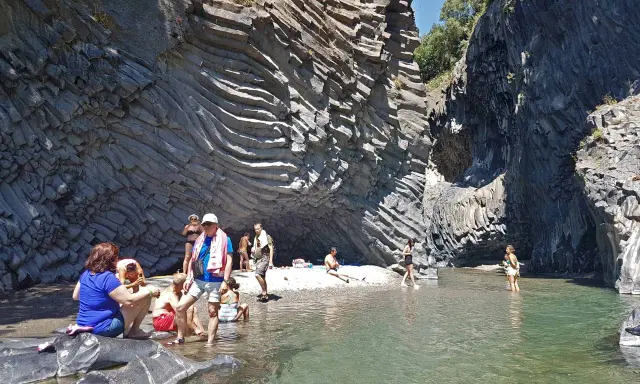 Persone si rilassano e si rinfrescano nelle acque cristalline delle cascate dell'Alcantara, circondate da imponenti pareti rocciose basaltiche.
