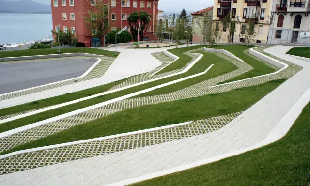 Diseño moderno con césped y caminos de hormigón que forman patrones ondulados. El suelo drenante permite el paso del agua, integrándose con el entorno urbano y el mar al fondo.