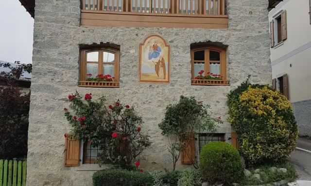 Un affresco sacro adorna un muro di pietra di un borgo trentino alto adige, con rose rosse che sbocciano rigogliose.