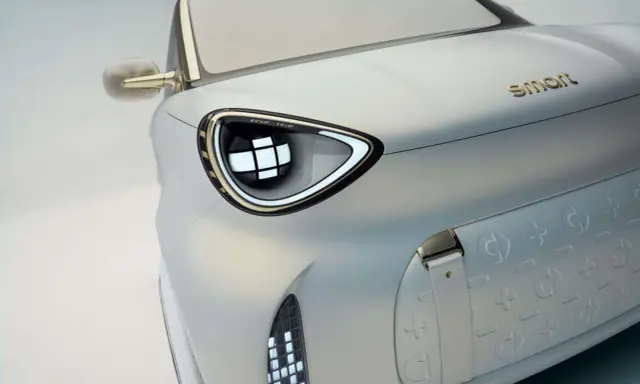 Dettaglio di un'auto elettrica Smart, con un faro futuristico e il logo dorato. Nuovi modelli in arrivo.