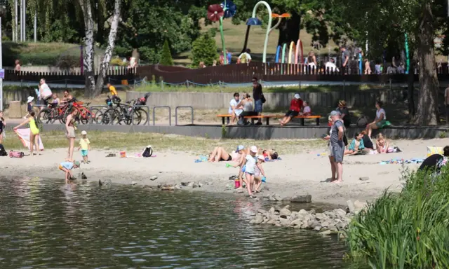 Rodzina odpoczywa nad wodą na piaszczystej plaży Jeziora Paprocany. Dzieci bawią się w piasku i przy brzegu, dorośli relaksują się na kocach i ławkach.