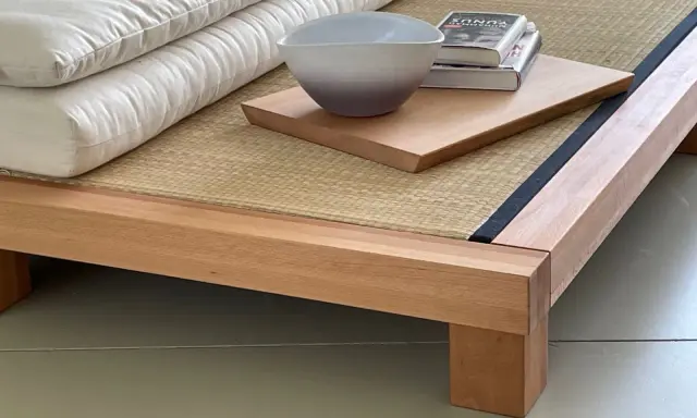 Eine gestapelte, weiße Matratze eines japanischen Bettes ruht auf einer Tatami-Matte. Daneben liegen ein Buch und eine Schale auf einem Holzbrett.
