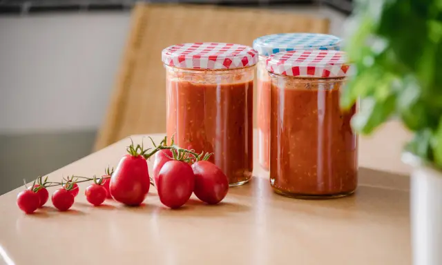 Frische Tomaten und eingekochte Tomatensauce in Gläsern. Die Frage "Tomatensauce einkochen wie lange?" beschäftigt viele Hobbyköche.
