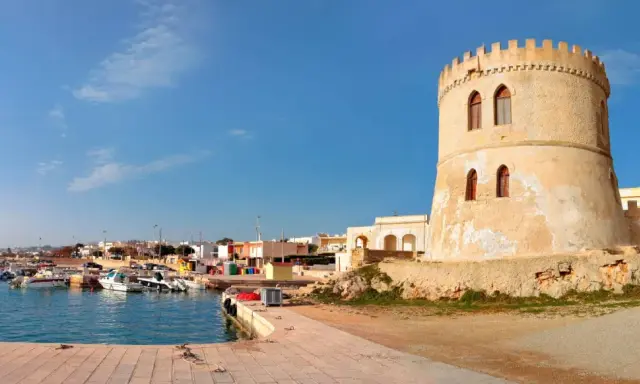 Torre Vado, Lecce: un'antica torre di avvistamento domina un porto con barche e case colorate sotto un cielo azzurro.