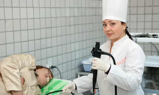 Lekarka w białym fartuchu i czepku przygotowuje pacjenta do gastroskopii, trzymając endoskop.