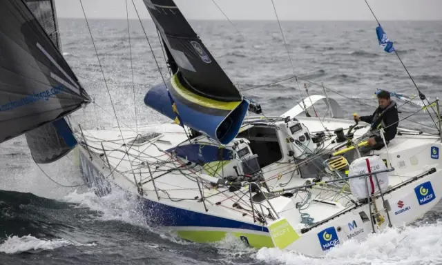 Un skipper navigue sur un voilier blanc et bleu, affrontant les vagues. Le nom "Pierre Quiroga" est visible sur la coque.