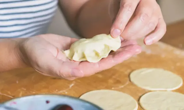 Dłonie formują ciasto na pierogi, tworząc pyszne danie. Na blacie leżą okrągłe placki z ciasta, a obok miseczka z wiśniami.
