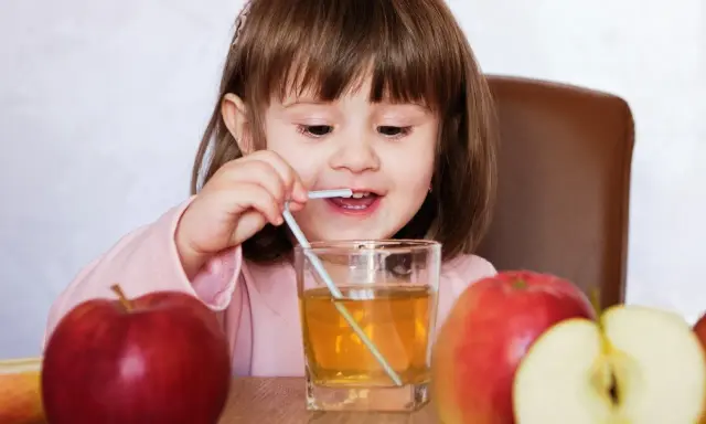 Mała dziewczynka pije sok jabłkowy, który pomaga na zaparcia. Obok stoją jabłka.