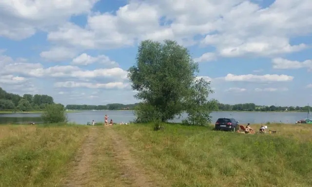 Słoneczny dzień nad jeziorem przeczyckim. Ludzie odpoczywają na trawie, w tle widać wodę i drzewa.