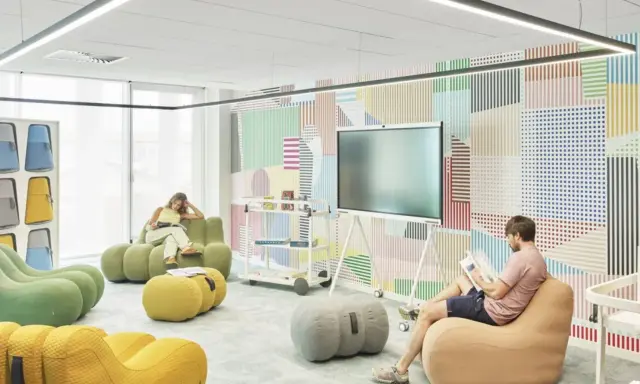 Un espace de repos coloré avec des poufs confortables et un mur à motifs géométriques. Deux personnes lisent tranquillement.