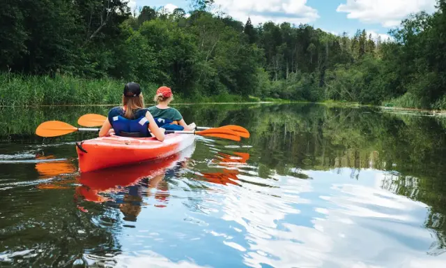Dwie osoby płyną pomarańczowym kajakiem po spokojnej rzece. Wokół bujna zieleń drzew i trzcin. Idealny spływ kajakowy Mazury.