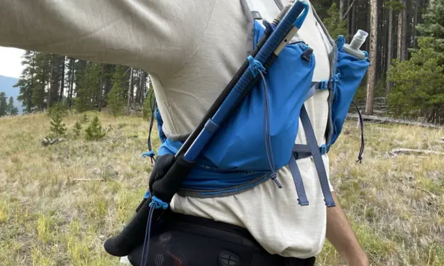 Jak przypiąć kijki do plecaka? Widok z boku na turystę w lesie, z kijami trekkingowymi przymocowanymi do niebieskiego plecaka.