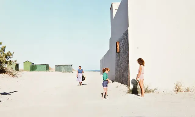 Luigi Ghirri foto: una donna cammina sulla spiaggia, due bambine giocano vicino a un edificio bianco.